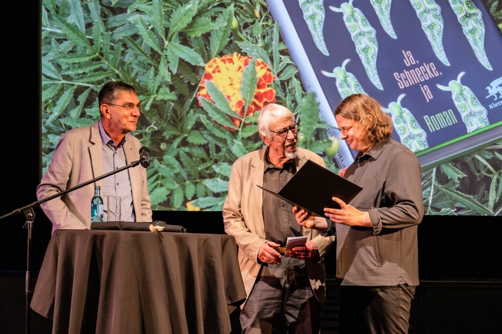 Auf der Bühne vor der Leinwand mit dem Buchcover von „Ja, Schnecke, ja“ reicht ein älterer Mann mit weißem Haar und Bart Jan Snela eine aufgeklappte Urkundenmappe. Jan Snela blickt lächelnd hinein. Links steht ein Mann im hellen Sakko an einem schwarz verhüllten Stehtisch.