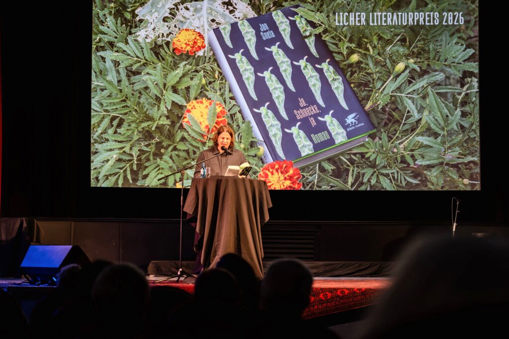 Jan Snela steht am Stehtisch auf der Bühne des Kino Traumstern und liest aus seinem Buch. Hinter ihm zeigt die Leinwand das Buchcover zwischen Studentenblumen und den Schriftzug „Licher Literaturpreis 2026“. Im Vordergrund die Köpfe des Publikums als Silhouetten.