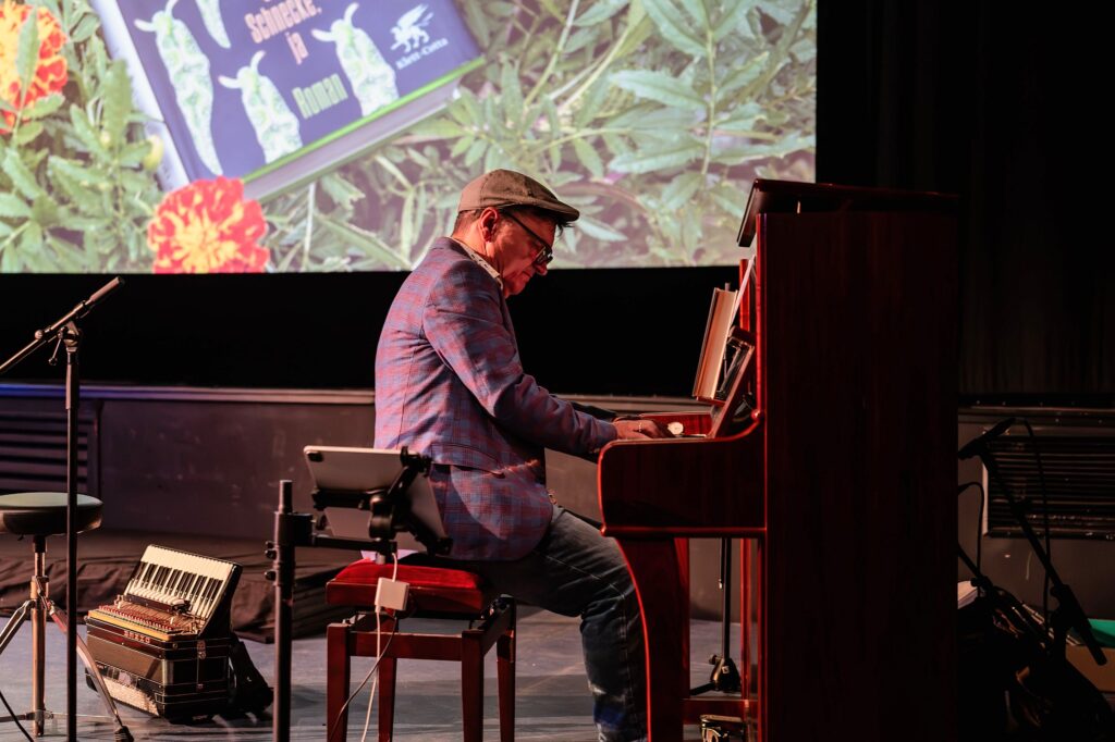 Helmut Fischer mit Schiebermütze und kariertem Sakko spielt Klavier. Neben ihm steht ein Akkordeon auf dem Bühnenboden, darüber ist ein Ausschnitt der Leinwand mit dem Buchcover zu sehen.